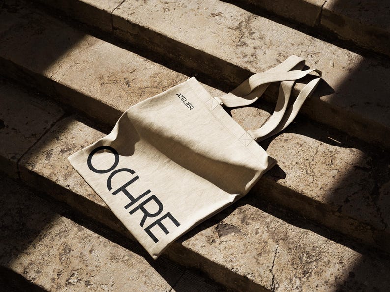 May include: A beige canvas tote bag with the word "OCHRE" printed in large, black capital letters. The word "ATELIER" is printed in smaller letters above the word "OCHRE". The bag has long, beige straps and is resting on stone steps.
