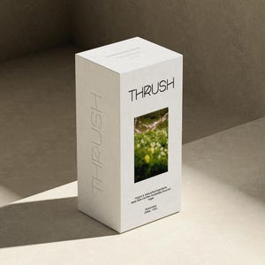 May include: A white box with the word "THRUSH" printed in a modern font. The box has a rectangular window displaying a green and yellow floral image. The word "THRUSH" is also embossed vertically on the side. The box is on a neutral surface.