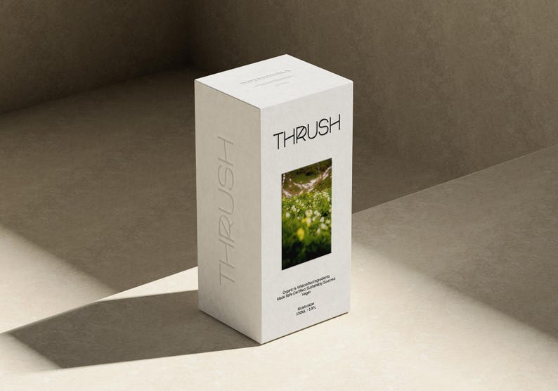 May include: A white box with the word "THRUSH" printed in a modern font. The box has a rectangular window displaying a green and yellow floral image. The word "THRUSH" is also embossed vertically on the side. The box is on a neutral surface.