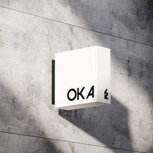 May include: A white, square sign with the word "OKA" in black, sans-serif font. The sign is mounted on a concrete wall. A black lightning bolt symbol is next to the letter "A".