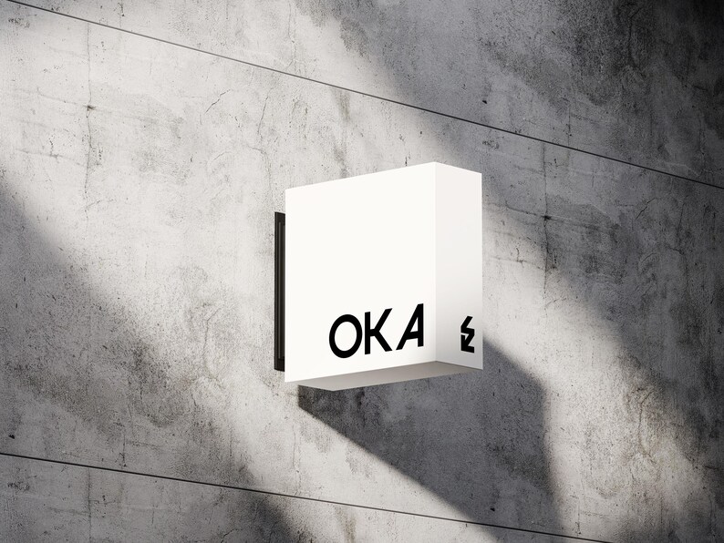 May include: A white, square sign with the word "OKA" in black, sans-serif font. The sign is mounted on a concrete wall. A black lightning bolt symbol is next to the letter "A".