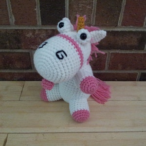 May include: A white crocheted unicorn with pink accents and large, black eyes. The unicorn has a gold horn and a pink tail.
