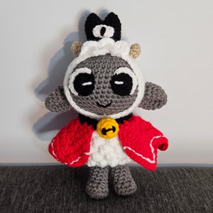 May include: Crocheted lamb plushie with a gray body, white face and collar, and a red cape. The lamb has large black eyes, a black crown, and a yellow bell. The handmade toy is a cute and unique gift.