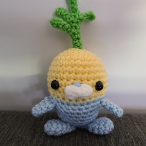 May include: A crocheted plush toy shaped like a turnip with a green stem, yellow body, and blue legs. The toy has black eyes and a white nose.