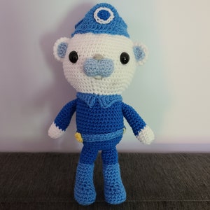 Puede incluir: Un oso de peluche de crochet que lleva un gorro azul y blanco con un círculo blanco. El oso lleva un atuendo de crochet azul con botones amarillos.