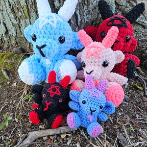 May include: Five crocheted plush toys in the shape of goats. The goats are in various colors, including blue, pink, black, and red. They are all wearing pentagram symbols on their chests. The goats are sitting on a bed of brown leaves and moss.