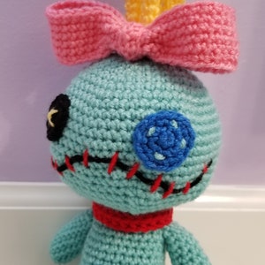 Puede incluir: Un juguete de peluche tejido a crochet que se asemeja a un personaje de la película de Disney Lilo & Stitch. El juguete es azul con un lazo rosa, un ojo negro, una boca roja y un botón azul.