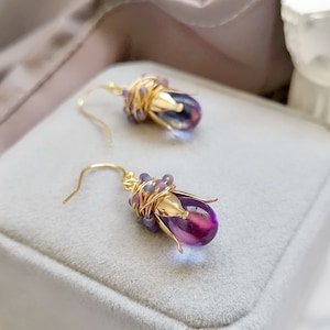 May include: A pair of purple gemstone earrings with gold wire wrapped around the stones and small purple beads. The earrings are displayed in a grey velvet box.