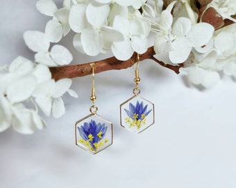 Pode incluir: Um par de brincos hexagonais dourados com um design de flores prensadas. As flores são azuis e amarelas e estão encapsuladas em resina transparente.