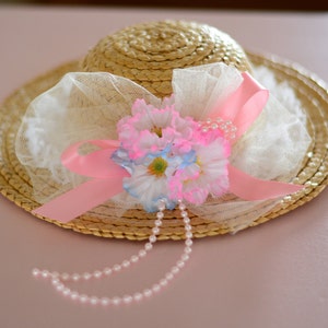 May include: A straw hat with a pink ribbon and white lace trim. The hat is decorated with pink and blue flowers and a pearl necklace.