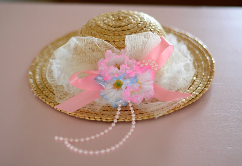May include: A straw hat with a pink ribbon and white lace trim. The hat is decorated with pink and blue flowers and a pearl necklace.
