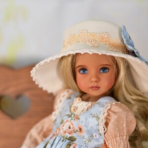 May include: A doll with blonde hair and blue eyes, wearing a light blue floral dress and a cream hat with lace trim. The hat has a blue ribbon. The doll has a fair complexion.