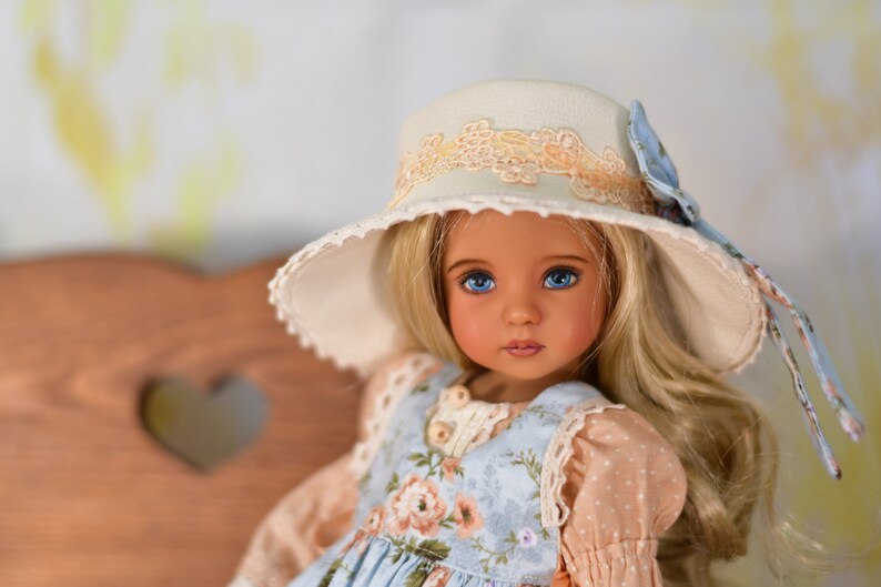 May include: A doll with blonde hair and blue eyes, wearing a light blue floral dress and a cream hat with lace trim. The hat has a blue ribbon. The doll has a fair complexion.