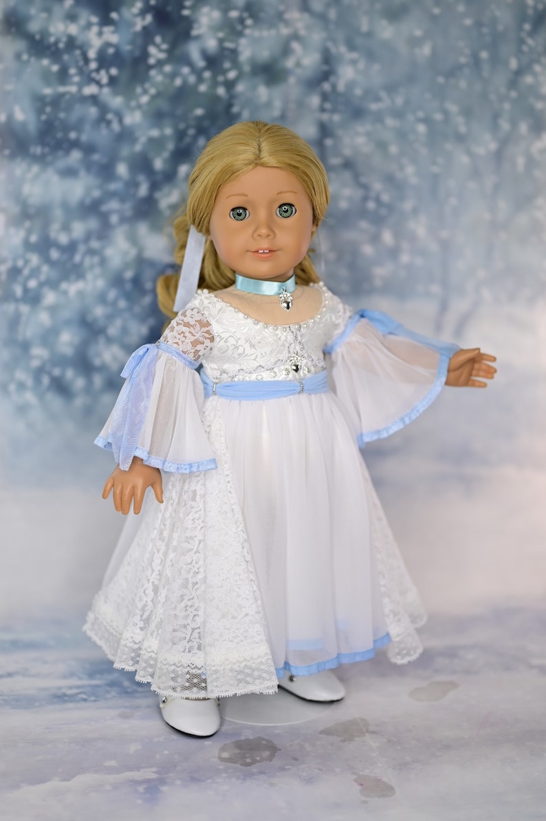 May include: A doll wearing a white lace dress with a light blue sash and matching collar. The doll has blonde hair and blue eyes. The background is a light blue with a snowy effect.