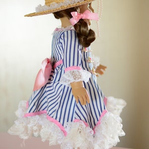 May include: A doll wearing a blue and white striped dress with a pink ribbon and lace trim. The doll is wearing a straw hat with a pink ribbon and a white lace trim. The doll is also wearing white boots with black accents.