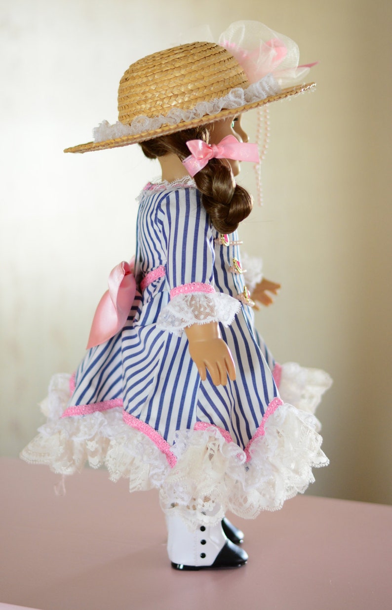 May include: A doll wearing a blue and white striped dress with a pink ribbon and lace trim. The doll is wearing a straw hat with a pink ribbon and a white lace trim. The doll is also wearing white boots with black accents.
