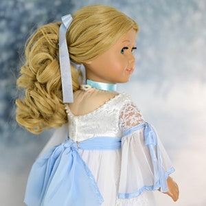 May include: A doll wearing a white dress with blue lace trim and a blue ribbon bow. The doll has blonde hair tied back in a ponytail.