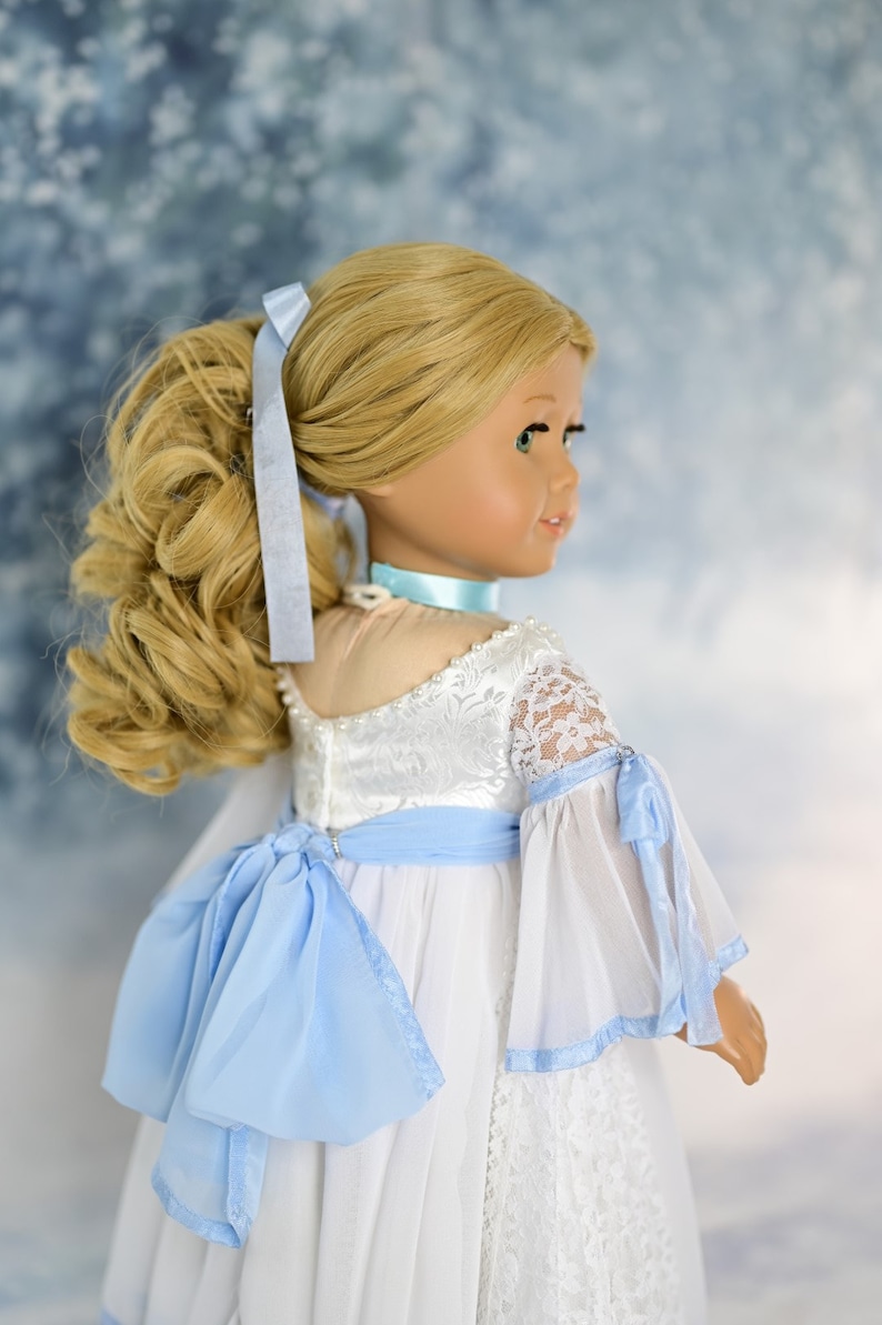 May include: A doll wearing a white dress with blue lace trim and a blue ribbon bow. The doll has blonde hair tied back in a ponytail.