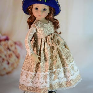 K&ouml;nnte beinhalten: Puppe mit blauem Hut mit Stoffblumen und einem beigefarbenen Kleid mit Blumenmuster. Das Kleid hat eine Spitzenborte und einen Bandsch&auml;rpe. Die Puppe tr&auml;gt schwarze Schuhe mit Schnallen.