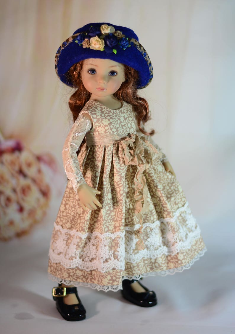 K&ouml;nnte beinhalten: Puppe mit blauem Hut mit Stoffblumen und einem beigefarbenen Kleid mit Blumenmuster. Das Kleid hat eine Spitzenborte und einen Bandsch&auml;rpe. Die Puppe tr&auml;gt schwarze Schuhe mit Schnallen.