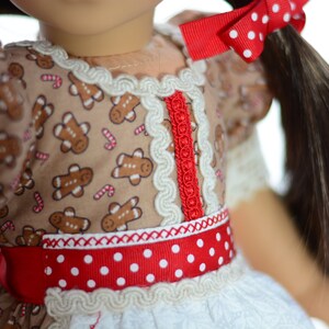 May include: A doll wearing a brown dress with gingerbread men and candy cane print. The dress has a red and white polka dot ribbon around the waist and white lace trim. The doll has brown hair and is wearing a red bow with white polka dots.