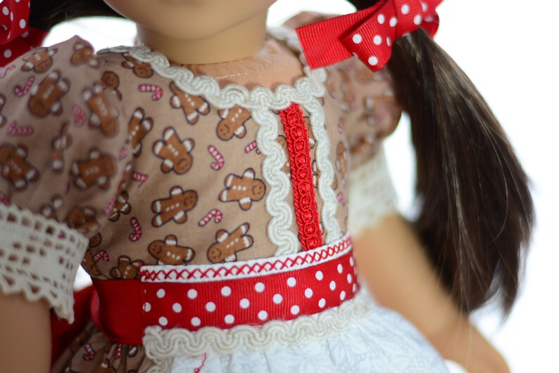May include: A doll wearing a brown dress with gingerbread men and candy cane print. The dress has a red and white polka dot ribbon around the waist and white lace trim. The doll has brown hair and is wearing a red bow with white polka dots.