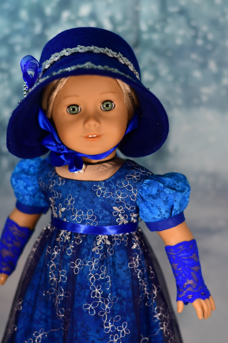 May include: A doll wearing a blue dress with silver floral embroidery, a blue hat with silver trim, and blue lace gloves.