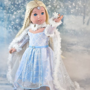 May include: A doll wearing a blue and white dress with a white fur-trimmed cape. The doll has long blonde hair and blue eyes. The doll is holding a small snowflake in her hand. The background is a snowy scene.