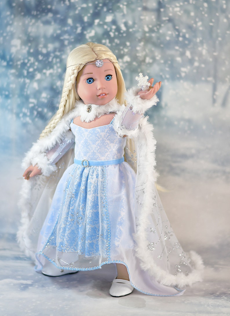 May include: A doll wearing a blue and white dress with a white fur-trimmed cape. The doll has long blonde hair and blue eyes. The doll is holding a small snowflake in her hand. The background is a snowy scene.