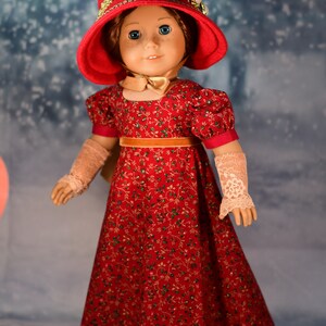 May include: A doll wearing a red dress with a floral pattern, a red hat with gold trim, and white lace gloves. The doll is standing on a snowy background.