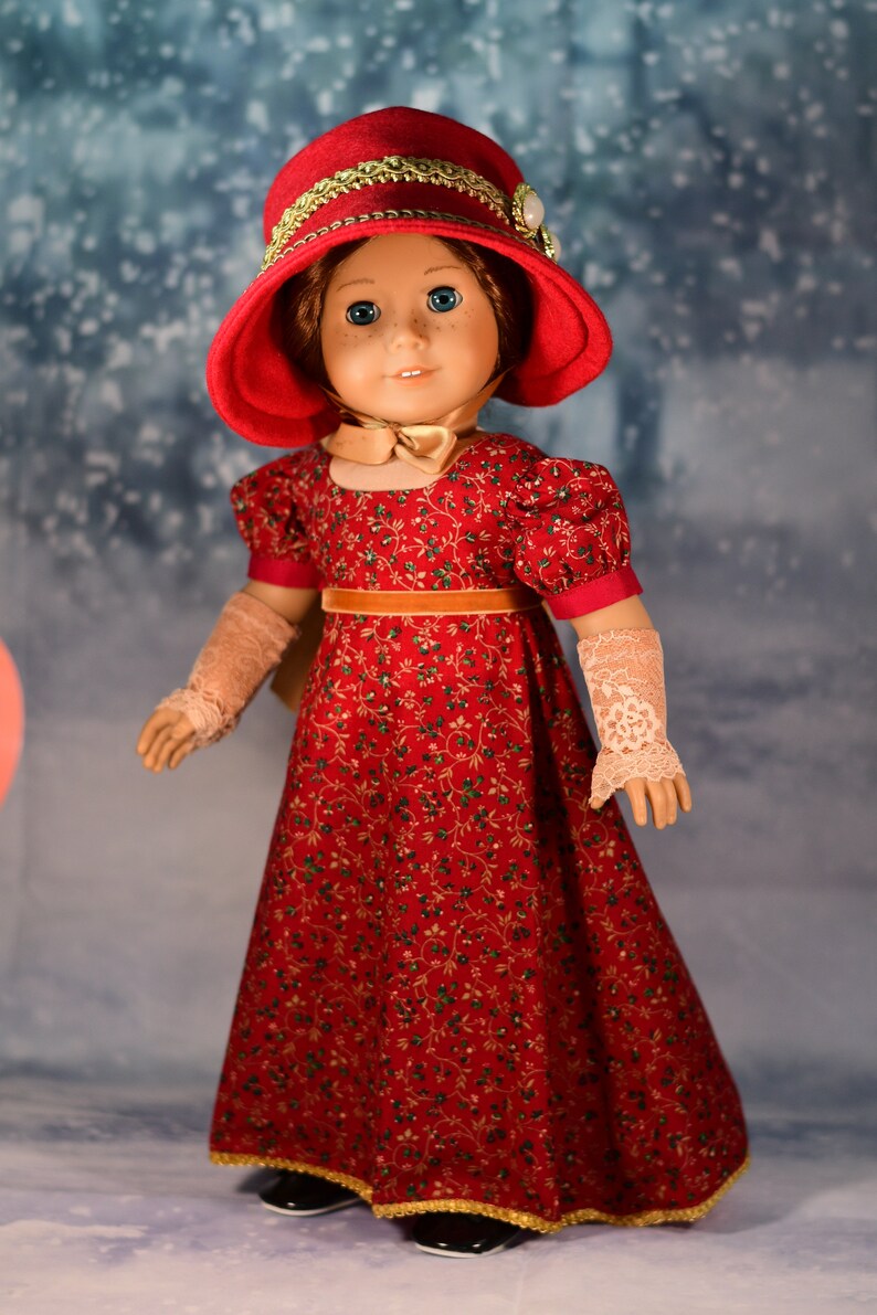 May include: A doll wearing a red dress with a floral pattern, a red hat with gold trim, and white lace gloves. The doll is standing on a snowy background.
