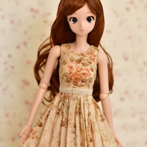 May include: A doll with long brown hair and large blue eyes, wearing a beige floral print dress with a lace waist. The dress features a rose pattern in shades of orange and green. The doll is posed against a soft floral background.