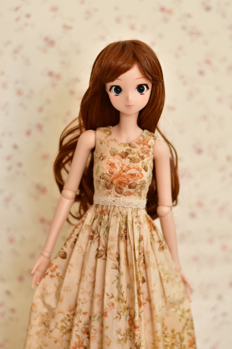 May include: A doll with long brown hair and large blue eyes, wearing a beige floral print dress with a lace waist. The dress features a rose pattern in shades of orange and green. The doll is posed against a soft floral background.