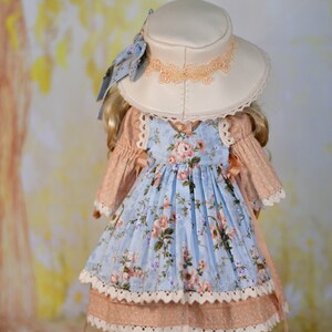May include: A doll dressed in a light blue floral dress and a matching hat. The dress features a pink bodice and white lace trim. The cream-coloured hat has peach-coloured lace. The doll is wearing white shoes.