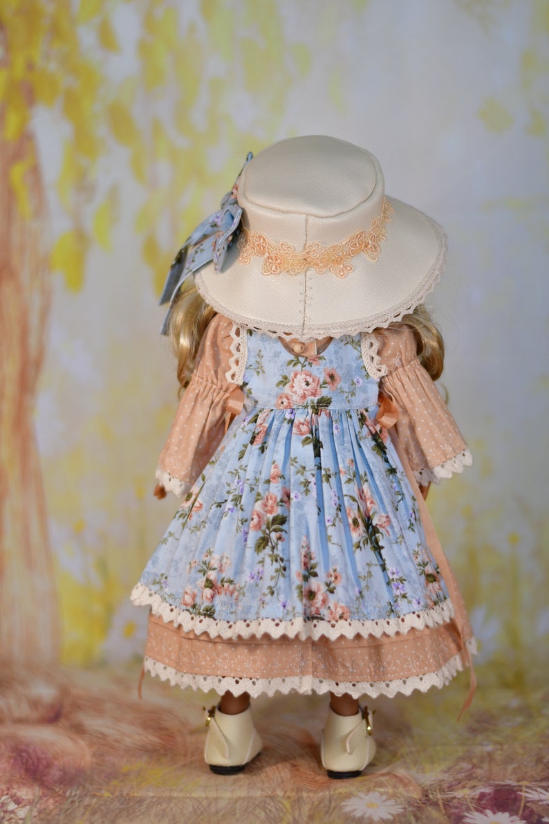 May include: A doll dressed in a light blue floral dress and a matching hat. The dress features a pink bodice and white lace trim. The cream-coloured hat has peach-coloured lace. The doll is wearing white shoes.