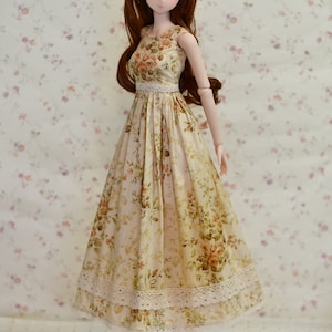 May include: A doll wearing a floral print dress with a white lace trim. The dress is beige with a pattern of orange and green flowers. The doll has long brown hair and is wearing brown shoes.