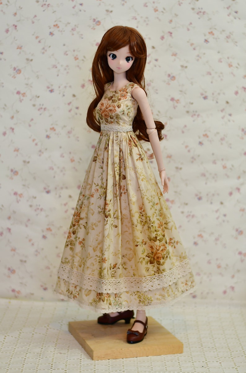 May include: A doll wearing a floral print dress with a white lace trim. The dress is beige with a pattern of orange and green flowers. The doll has long brown hair and is wearing brown shoes.