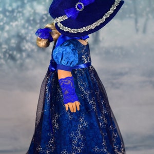 May include: A blue and silver doll's dress with a matching blue hat and gloves. The dress has a lace overlay and a satin ribbon sash. The hat has a silver trim and a blue bow.