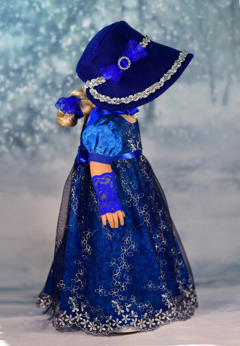 May include: A blue and silver doll's dress with a matching blue hat and gloves. The dress has a lace overlay and a satin ribbon sash. The hat has a silver trim and a blue bow.