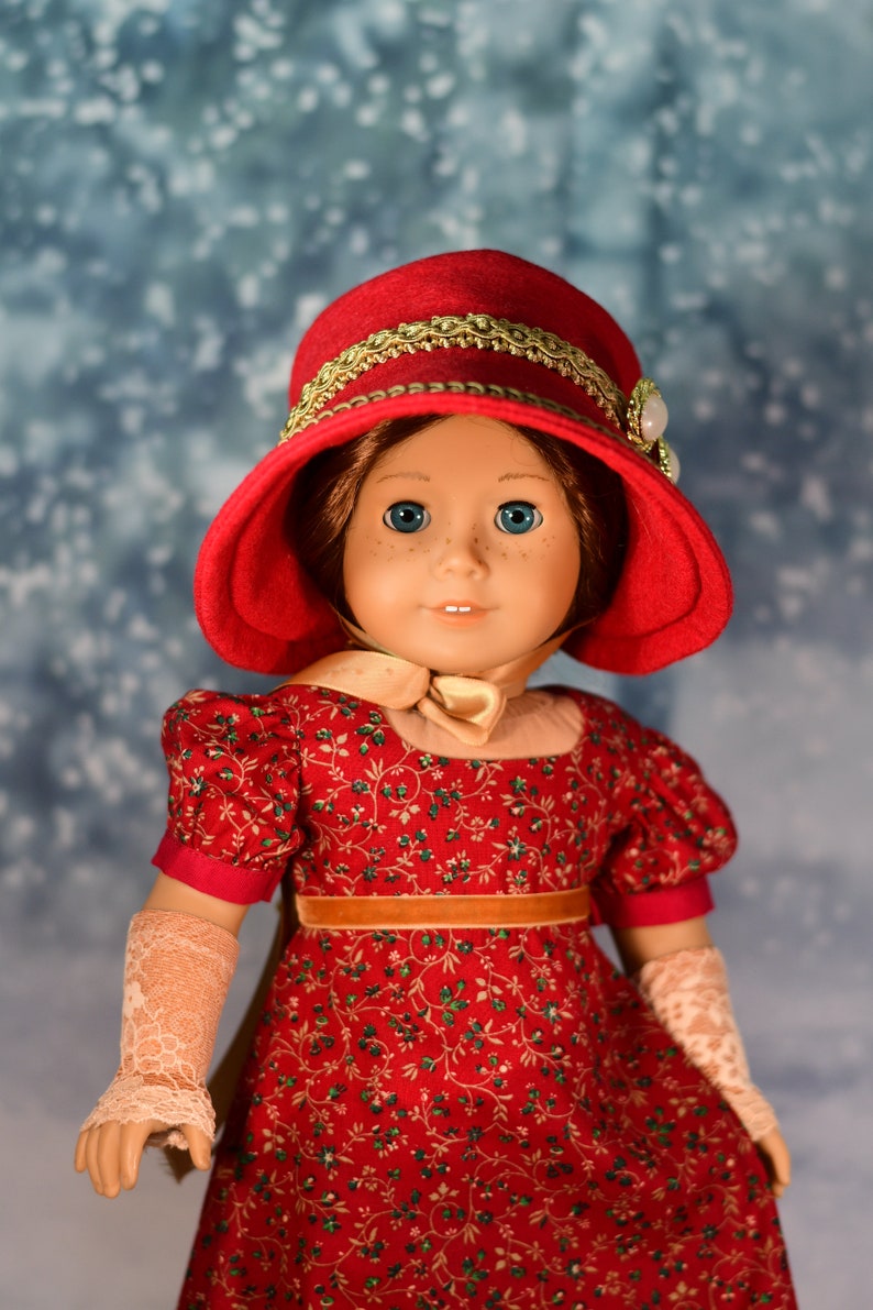 May include: A red doll wearing a red hat with gold trim and a red and green floral dress. The doll has blue eyes and brown hair.