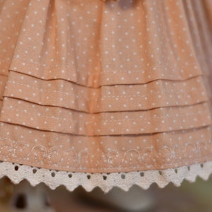 May include: Close-up of a peach fabric with white polka dots, featuring layered ruffles. The bottom edge has a delicate white lace trim with a scalloped design. The fabric appears to be part of a dress or skirt.