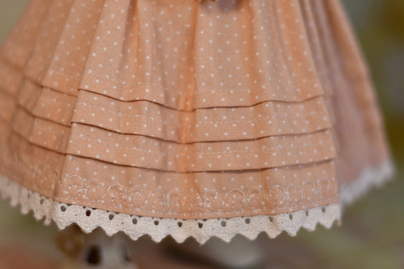 May include: Close-up of a peach fabric with white polka dots, featuring layered ruffles. The bottom edge has a delicate white lace trim with a scalloped design. The fabric appears to be part of a dress or skirt.