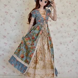 May include: A doll wearing a vintage-style dress with a floral pattern in shades of blue, cream, and red. The dress features lace trim and a matching hat. The doll is standing on a wooden base.