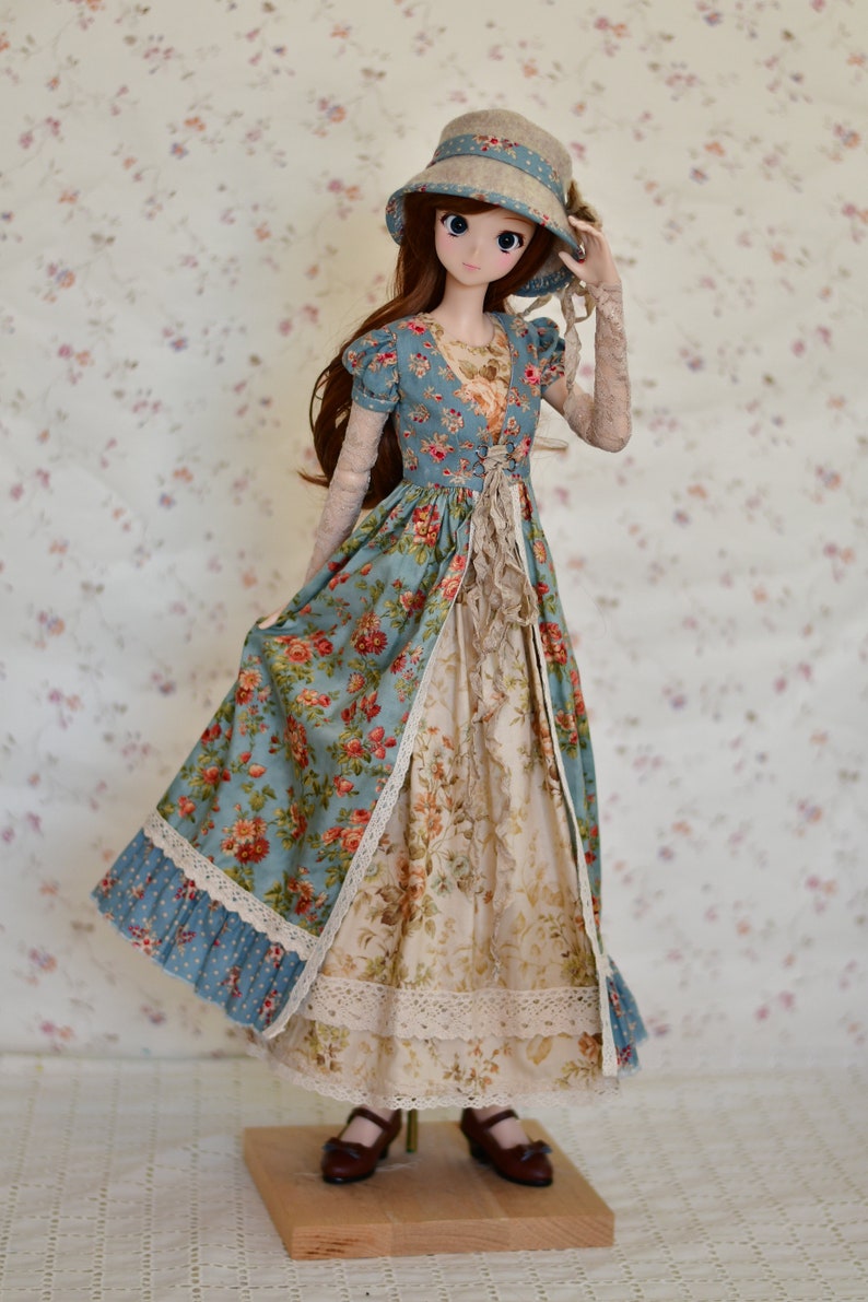 May include: A doll wearing a vintage-style dress with a floral pattern in shades of blue, cream, and red. The dress features lace trim and a matching hat. The doll is standing on a wooden base.