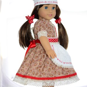 May include: A doll wearing a brown and white gingerbread-themed dress with a red ribbon and white apron. The doll is wearing a white chef's hat with red trim and has brown hair in pigtails with red polka dot bows.