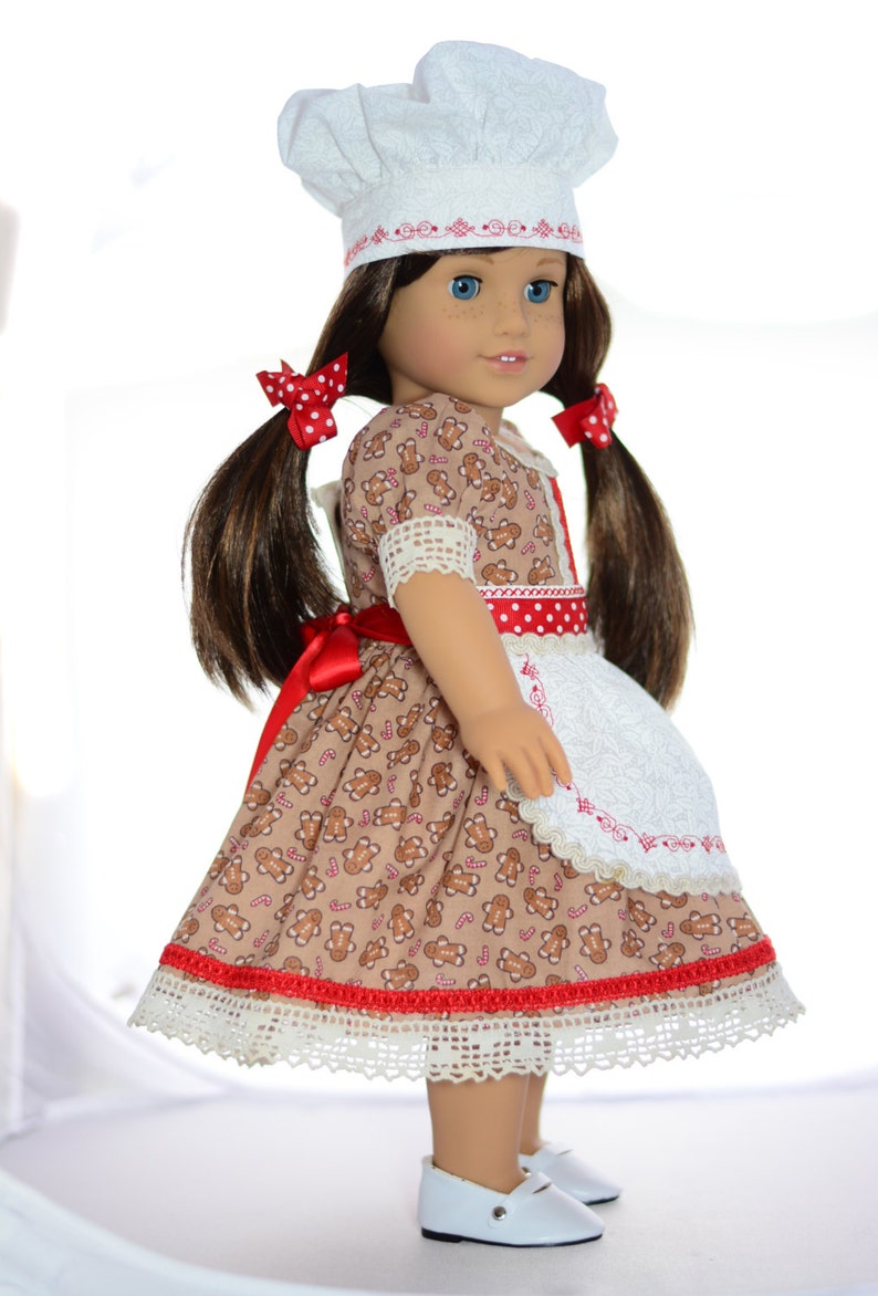 May include: A doll wearing a brown and white gingerbread-themed dress with a red ribbon and white apron. The doll is wearing a white chef's hat with red trim and has brown hair in pigtails with red polka dot bows.
