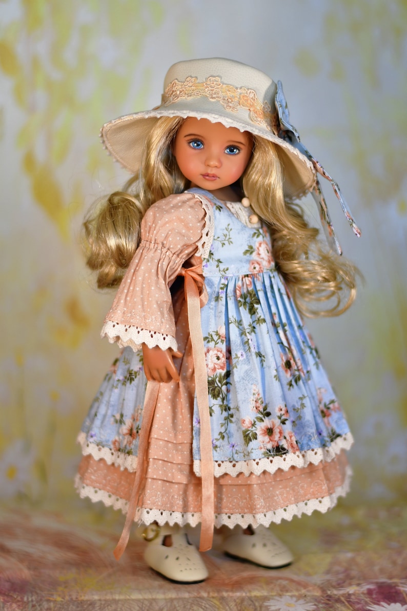 May include: A doll in a light blue floral dress with a peach sleeve and trim. The doll has blonde hair and a matching hat with a floral bow. The doll is wearing white shoes.