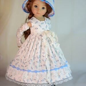 K&ouml;nnte beinhalten: Puppe in einem wei&szlig;en Kleid mit blauem Blumenmuster, Spitzensaum und blauen Banddetails. Die Puppe tr&auml;gt auch einen passenden Hut mit floralen Akzenten und schwarzen Schuhen.