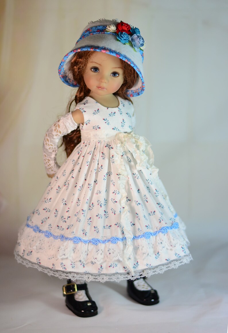 K&ouml;nnte beinhalten: Puppe in einem wei&szlig;en Kleid mit blauem Blumenmuster, Spitzensaum und blauen Banddetails. Die Puppe tr&auml;gt auch einen passenden Hut mit floralen Akzenten und schwarzen Schuhen.
