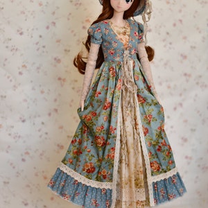 May include: A doll wearing a blue floral print dress with a matching hat. The dress features a V-neck, lace trim, and a ruffled front. The doll has long brown hair and is wearing brown shoes. The background is a floral pattern.